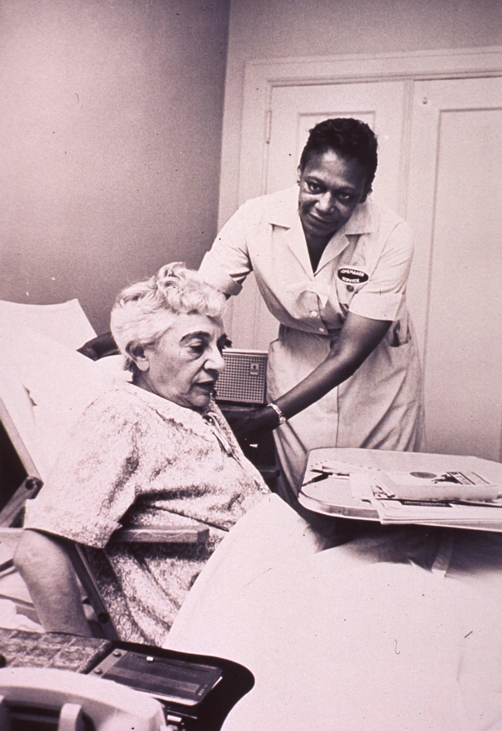 Caregiver in uniform assists elderly woman sitting in bed.